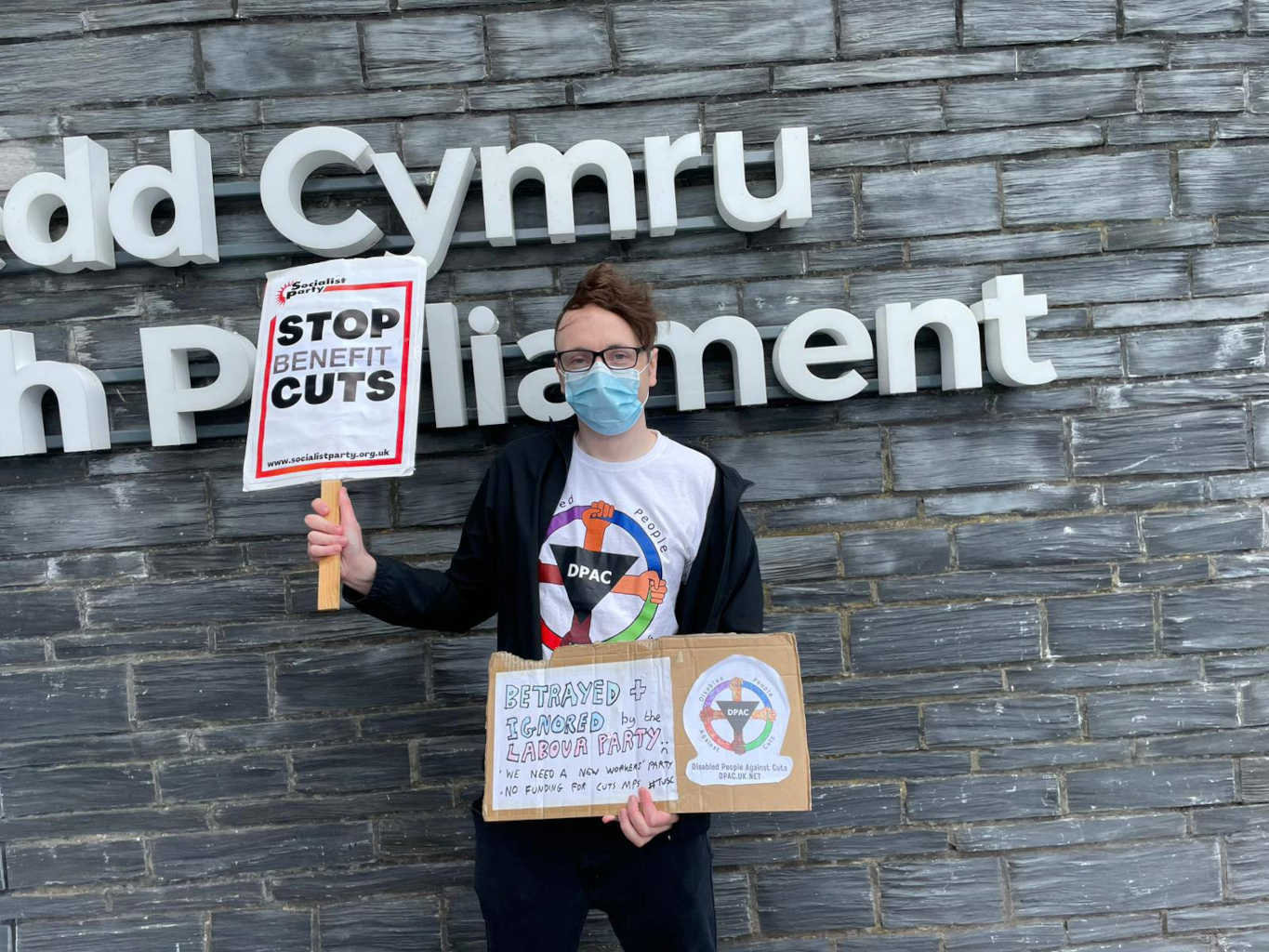 Ben Golightly at the Senedd. He is wearing a Disabled People Against Cuts (DPAC) shirt, carrying a placard that says 'Stop Benefit Cuts', and a homemade placard that says 'betrayed and ignored by the Labour Party'.