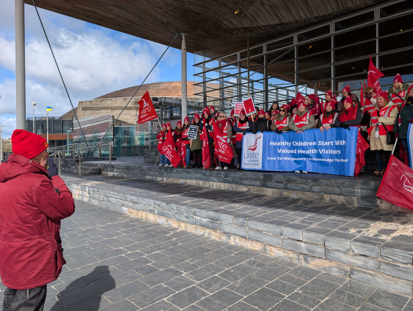 A man in red is speaking on a megaphone to a crowd. They are outside the Senedd. The crowd has blue banners and red flags.