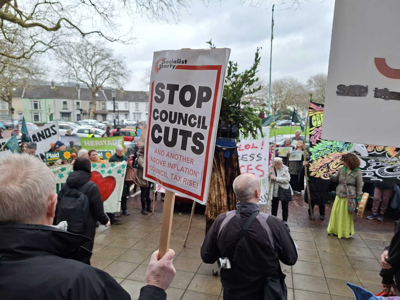 At a colourful protest in Swansea, a man in the foreground is holding a Socialist Party placard that says 'stop council cuts.'